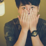 A surprised young man covers his face with his hands wearing a camouflage shirt indoors.